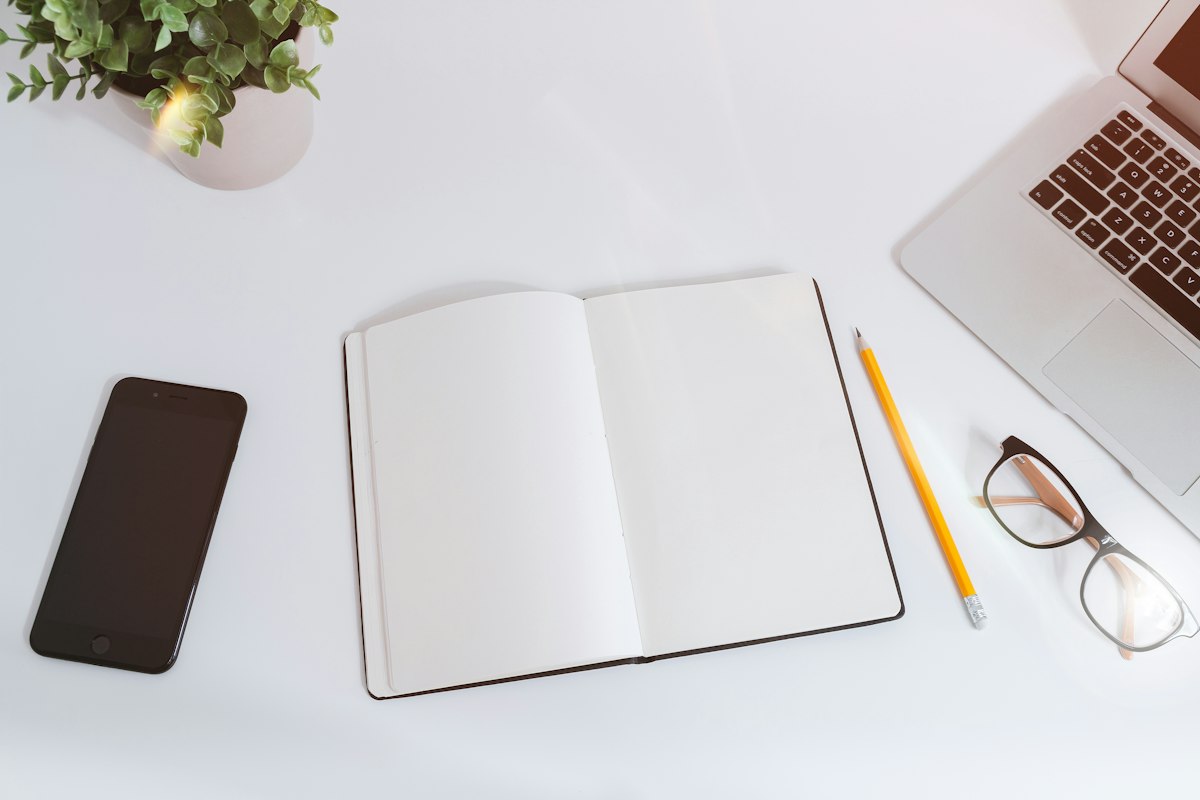 An overhead shot of a tidy white desk with a laptop, a smartphone, glasses, an open notebook, and a pencil — generic workspace stock photo illustrating the mail-and-calendar-client section below, not the Penguin Mail interface or its AI assistant panel