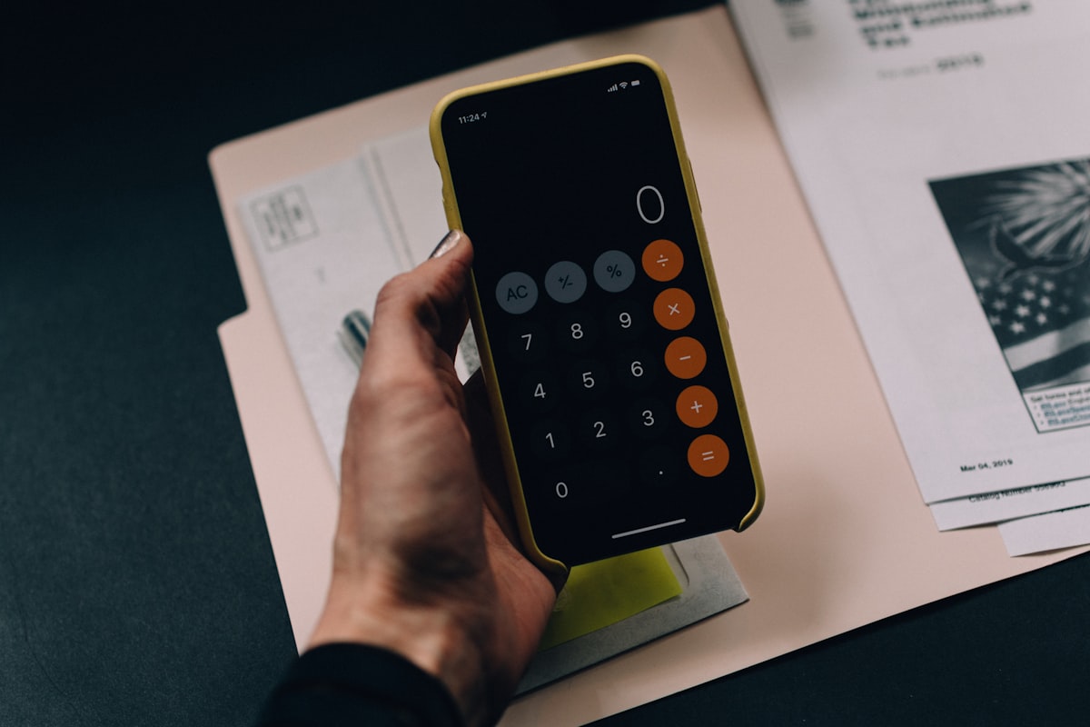 Hand holding a smartphone calculator app over financial documents — a generic business technology image unrelated to actual stock chart data